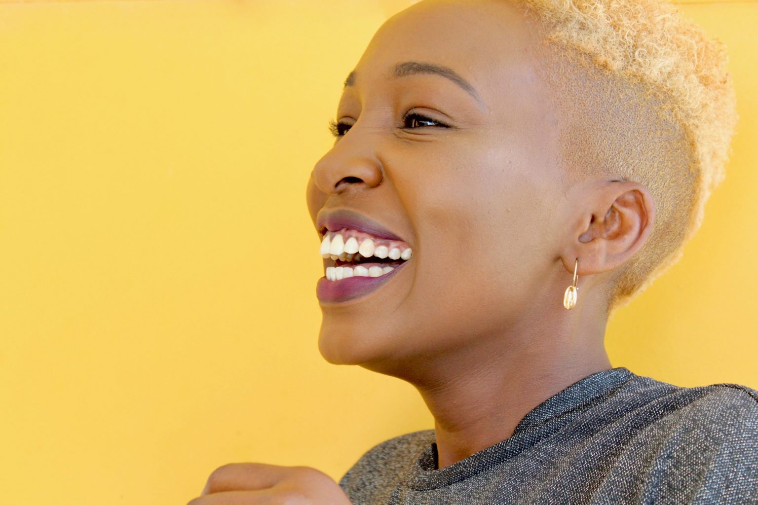 A black woman with short hair and a gummy smile on a bright yellow background