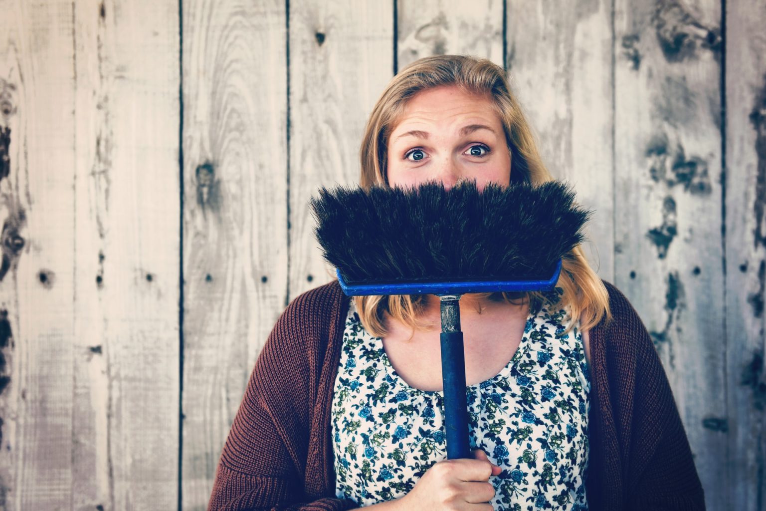 A young, blonde woman hides her smile behind a broom